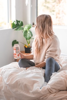 A person with long blonde hair sits on a bed holding a carton labeled 'Boxed Water is Better.' The person is dressed in a cozy sweater and leggings. Behind them, a large window allows natural light to fill the room, and a potted plant with broad leaves is visible in the corner.