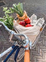 A bicycle handlebar and basket filled with fresh groceries, including leeks, apples, leafy greens, and boxed water. The bike is on a cobblestone surface and has a silver bell with branding on it.