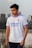 A person is standing outdoors wearing a white sports jersey with blue and orange accents. The jersey has brand logos and text on it. The background suggests an urban setting with buildings and a cloudy sky.