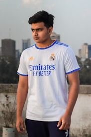 A person is standing outdoors wearing a white sports jersey with blue and orange accents. The jersey has brand logos and text on it. The background suggests an urban setting with buildings and a cloudy sky.