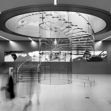 A modern spiral staircase made of glass and metal dominates the center of a sleek, futuristic interior. The staircase is enclosed in a transparent cylindrical structure, with a minimalist design that is both functional and artistic. The lighting is integrated into the ceiling, providing an even illumination. Shadows and reflections create a dynamic interplay on the walls and floor. A mural on the wall adds an abstract artistic touch to the space.