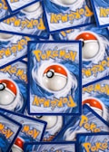 Multiple Pokémon trading cards are scattered, with the back side visible. Each card features a large red and white Pokéball design in the center, surrounded by swirling blue patterns and prominently displaying the Pokémon logo in bright yellow lettering.
