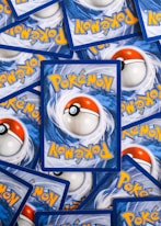Multiple Pokémon trading cards are scattered, with the back side visible. Each card features a large red and white Pokéball design in the center, surrounded by swirling blue patterns and prominently displaying the Pokémon logo in bright yellow lettering.