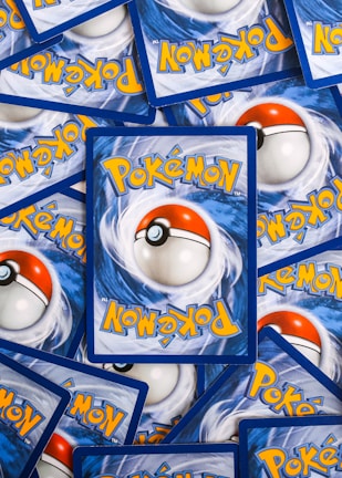 Multiple Pokémon trading cards are scattered, with the back side visible. Each card features a large red and white Pokéball design in the center, surrounded by swirling blue patterns and prominently displaying the Pokémon logo in bright yellow lettering.