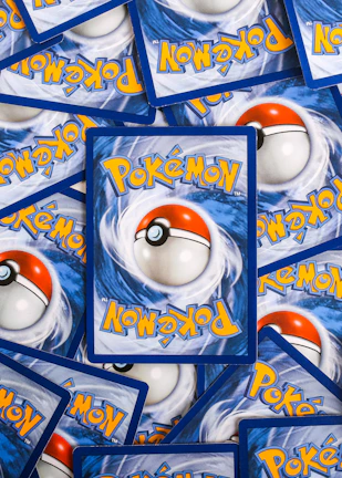Multiple Pokémon trading cards are scattered, with the back side visible. Each card features a large red and white Pokéball design in the center, surrounded by swirling blue patterns and prominently displaying the Pokémon logo in bright yellow lettering.