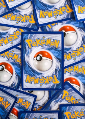 Multiple Pok&eacute;mon trading cards are scattered, with the back side visible. Each card features a large red and white Pok&eacute;ball design in the center, surrounded by swirling blue patterns and prominently displaying the Pok&eacute;mon logo in bright yellow lettering.