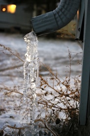 A frozen gutter downspout with icicles hanging from it, set against a snowy background with dry grass and a blurred house in the distance.