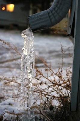 A frozen gutter downspout with icicles hanging from it, set against a snowy background with dry grass and a blurred house in the distance.
