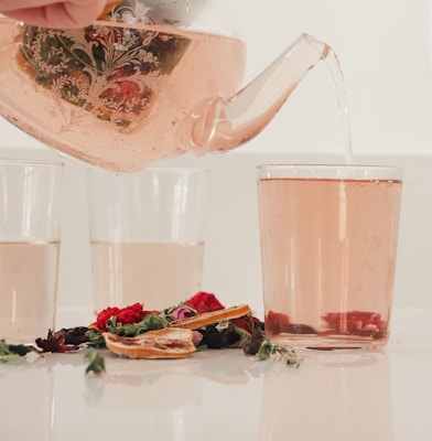 A glass teapot with intricate floral designs is pouring a light pink liquid into a glass. Several other glasses contain the same pink liquid. In the foreground, there are dried flower petals and slices of dried citrus fruit scattered on a reflective surface.