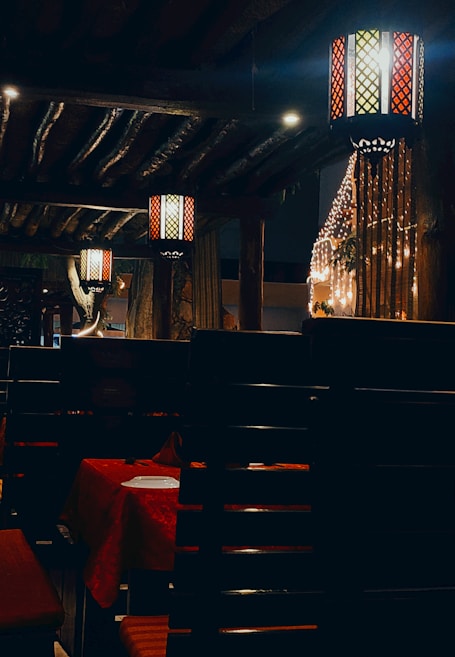 The image depicts a dimly lit interior space, possibly a restaurant. Ornate lanterns with colorful glass panels hang from the wooden ceiling, casting warm light throughout the room. The setting includes dark wooden furniture and tables covered with red tablecloths. Strings of decorative lights add an atmospheric touch.