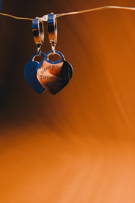 A pair of heart-shaped silver pendants hang elegantly from a wire against a rich, warm brown background. The pendants are engraved with the text 'PLEASE RETURN TO' and 'TIFFANY & CO.' along with other details. The lighting highlights the reflective surface of the silver, creating a luxurious and sophisticated ambiance.
