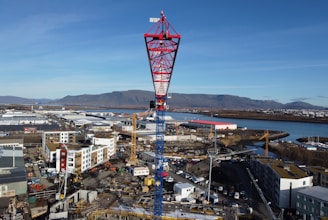 A large red and blue construction crane overlooks an extensive construction site filled with various buildings and equipment. In the background, the landscape includes a river, open fields, industrial buildings, and distant mountains under a clear blue sky.