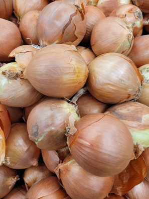 A large pile of brown onions with papery skins, some of which have slight tears. The onions are close together, showcasing their round shapes and the texture of their outer layers.