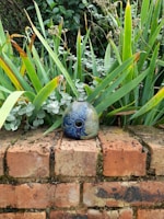 A decorative ceramic object with intricate floral patterns is placed on a weathered brick wall. Surrounded by lush green foliage, the leaves show a mix of vibrant and dried tips, creating a contrast against the detailed blue and green hues of the ceramic.
