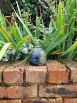 A decorative ceramic object with intricate floral patterns is placed on a weathered brick wall. Surrounded by lush green foliage, the leaves show a mix of vibrant and dried tips, creating a contrast against the detailed blue and green hues of the ceramic.