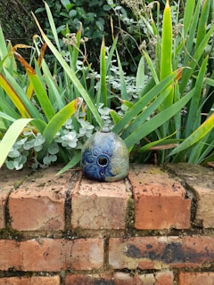 A decorative ceramic object with intricate floral patterns is placed on a weathered brick wall. Surrounded by lush green foliage, the leaves show a mix of vibrant and dried tips, creating a contrast against the detailed blue and green hues of the ceramic.