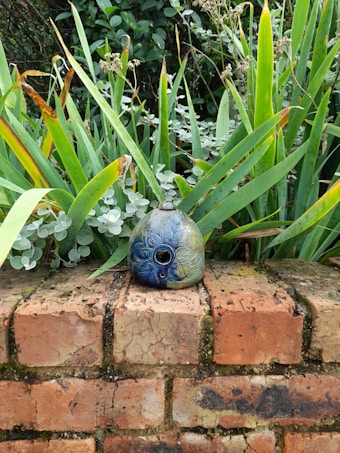 A decorative ceramic object with intricate floral patterns is placed on a weathered brick wall. Surrounded by lush green foliage, the leaves show a mix of vibrant and dried tips, creating a contrast against the detailed blue and green hues of the ceramic.