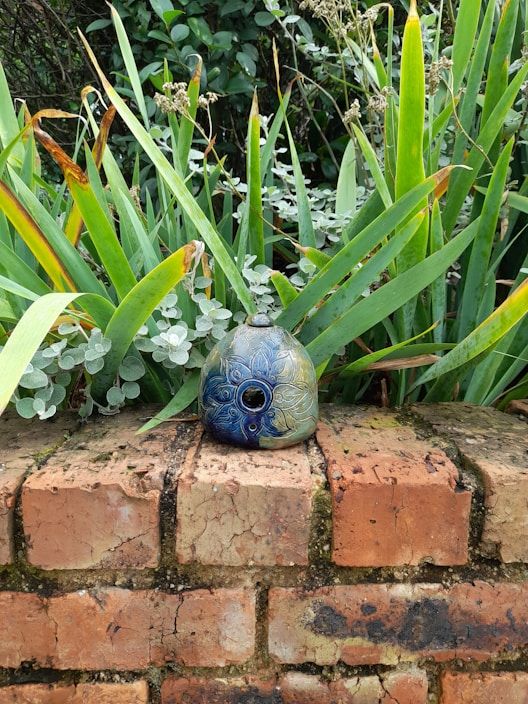 A decorative ceramic object with intricate floral patterns is placed on a weathered brick wall. Surrounded by lush green foliage, the leaves show a mix of vibrant and dried tips, creating a contrast against the detailed blue and green hues of the ceramic.