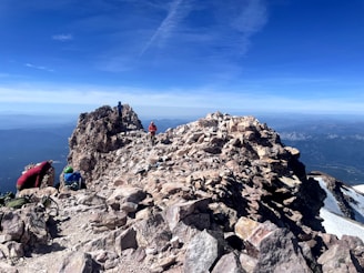 A rugged, rocky mountain peak with climbers ascending the trail. Several people are visible, wearing bright outdoor gear. The sky is clear and blue, offering a breathtaking view of distant mountain ranges and valleys.