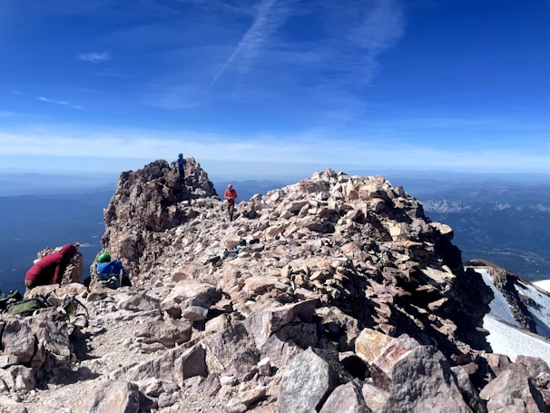 A rugged, rocky mountain peak with climbers ascending the trail. Several people are visible, wearing bright outdoor gear. The sky is clear and blue, offering a breathtaking view of distant mountain ranges and valleys.