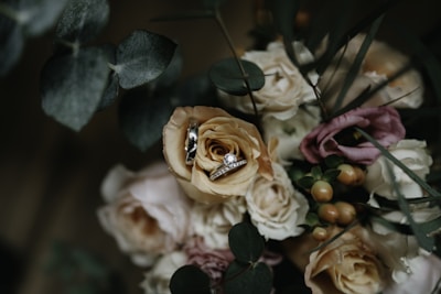 A delicate arrangement of roses and other flowers, with a set of wedding rings placed atop a peach-colored rose. The floral arrangement includes green leaves and berries, creating a lush and romantic setting.