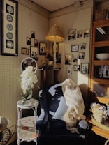 A cozy corner featuring a dark upholstered chair adorned with a light-colored knitted throw and a cushion. A small side table with white flowers in a vase stands beside the chair. Numerous framed black and white photos and artwork hang neatly on the pegboard walls. A rattan lampshade casts a warm light, contributing to the vintage aesthetic of the space.