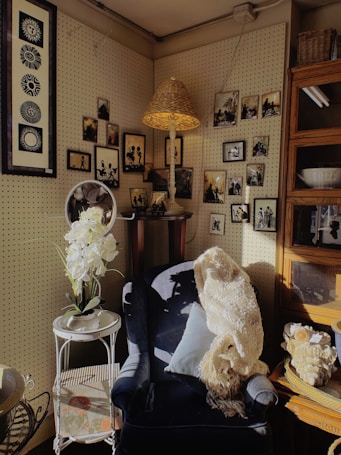 A cozy corner featuring a dark upholstered chair adorned with a light-colored knitted throw and a cushion. A small side table with white flowers in a vase stands beside the chair. Numerous framed black and white photos and artwork hang neatly on the pegboard walls. A rattan lampshade casts a warm light, contributing to the vintage aesthetic of the space.