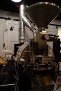 A large industrial coffee roasting machine with a metal hopper at the top, a gauge, and various pipes and levers. The machine is situated inside a workshop with ducts and tools in the background.