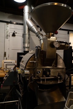 A large industrial coffee roasting machine with a metal hopper at the top, a gauge, and various pipes and levers. The machine is situated inside a workshop with ducts and tools in the background.