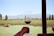 A person holds a glass of red wine overlooking a vineyard with rows of grapevines. In the background, there are tall green trees and distant mountains under a clear blue sky. The scene is framed by a wooden post on the right, suggesting an outdoor terrace or patio.