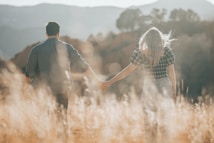 A couple is walking hand in hand through a sunlit field, surrounded by tall grasses. The scene is set against a backdrop of hills and softly blurred trees, creating a dreamy atmosphere. The lighting is warm, with the sun casting a gentle glow around them.