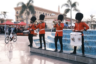 Four musicians dressed in traditional British royal guard uniforms are standing by a fountain, playing instruments like the trombone, trumpet, snare drum, and bass drum. There are people in the background, including a child on a bicycle. The setting appears to be a festive outdoor area with tents and palm trees.