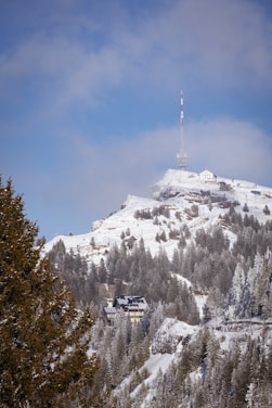 A snow-covered mountain landscape featuring evergreen trees and a few buildings nestled among the trees. Atop the mountain stands a tall communication tower with red and white stripes, and the sky above is partly cloudy with patches of blue visible.