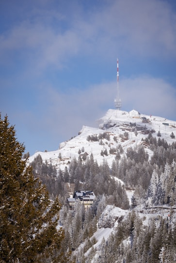 A snow-covered mountain landscape featuring evergreen trees and a few buildings nestled among the trees. Atop the mountain stands a tall communication tower with red and white stripes, and the sky above is partly cloudy with patches of blue visible.
