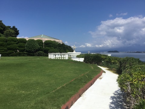 A well-maintained garden area with lush green grass and neatly trimmed bushes. A white pathway curves through the garden, leading towards a white decorative structure resembling a fountain. In the background, a building with a green roof is partially visible. The scene is set against a backdrop of a calm sea under a sky filled with some clouds.