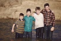 Four boys are standing in shallow water with their arms around each other, all wearing casual shirts and jeans. They appear happy and relaxed, with close smiles, suggesting camaraderie among them. The background shows a blurred, natural setting with earthy tones.