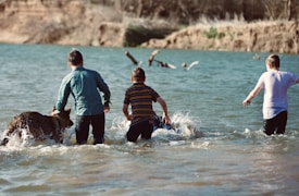 Three children are playing in the water with a dog at a lake. The scene is set in a natural environment with a view of the distant bank and trees. The children appear to be splashing and moving energetically through the shallow water.