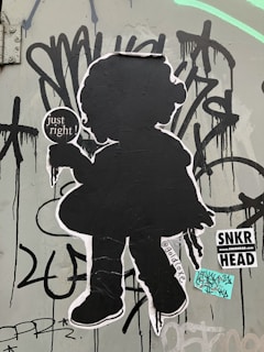 A black silhouette of a child holding an ice cream cone is pasted on a wall covered with graffiti. The silhouette includes the phrase 'just right!' and a social media handle '@goldoxc' on it. The wall is adorned with various graffiti tags and street art stickers, one of which reads 'SNKR HEAD'.