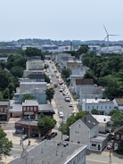 A suburban neighborhood with a mix of residential houses and small commercial buildings. A road runs through the center lined with parked cars. Trees and greenery are present throughout the area. In the distance, a large wind turbine and industrial buildings are visible.