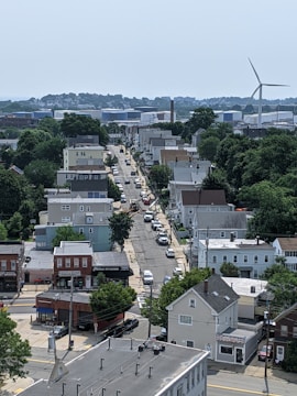 A suburban neighborhood with a mix of residential houses and small commercial buildings. A road runs through the center lined with parked cars. Trees and greenery are present throughout the area. In the distance, a large wind turbine and industrial buildings are visible.