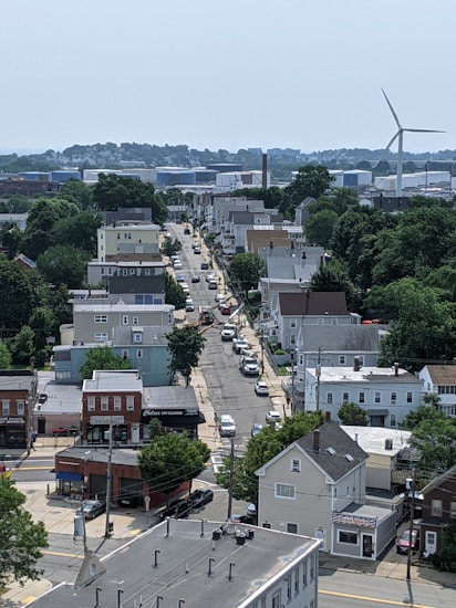 A suburban neighborhood with a mix of residential houses and small commercial buildings. A road runs through the center lined with parked cars. Trees and greenery are present throughout the area. In the distance, a large wind turbine and industrial buildings are visible.