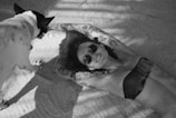 A person with tattoos lies on their back, wearing sunglasses and a bikini, on a sandy surface. A small dog with a short coat approaches, casting shadows on the sand. The scene is in black and white.