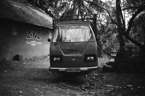 A black and white photograph of a three-wheeled vehicle parked on a dirt path. The vehicle appears to be stationary and is facing the camera, with a visible license plate and the word 'GOODS' on the front. The background includes a wall with the painted text 'ZINZi find any reason'. The scene is surrounded by trees and foliage, creating a rustic atmosphere.
