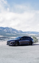 A sleek, dark-colored car is parked on a gravel area with a beautiful, vast mountainous landscape in the background. The image features clear blue skies with a few clouds, providing an expansive view of the mountains and valleys.