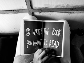 A person is holding an open book with the text '3) WRITE THE BOOK YOU WANT TO READ' written on one of the pages. The image is in black and white, showing a close-up view from above.