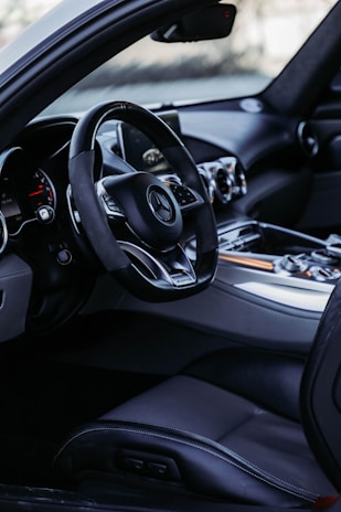 The interior of a luxury car featuring a sleek dashboard with multiple circular air vents, a leather-wrapped steering wheel with a prominent logo in the center, and a digital display screen. The seating is upholstered in high-quality black leather with visible stitching.