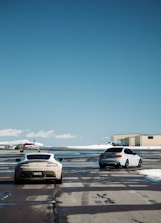Two cars are parked on an airport runway with a small airplane in the background. The cars are positioned on the tarmac next to each other, and a beige hangar is visible on the right. The ground is wet, reflecting the vehicles and part of the aircraft. The sky is clear and blue, indicating a sunny day.