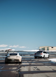 Two cars are parked on an airport runway with a small airplane in the background. The cars are positioned on the tarmac next to each other, and a beige hangar is visible on the right. The ground is wet, reflecting the vehicles and part of the aircraft. The sky is clear and blue, indicating a sunny day.