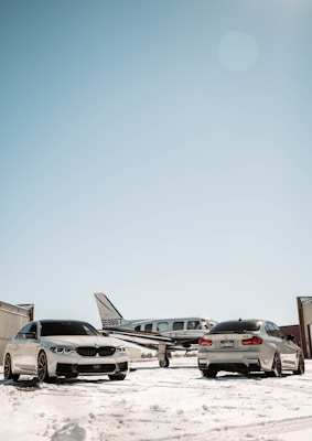 Two luxury cars are parked on a snow-covered surface in front of a small airplane at an airfield. The scene is set under a clear blue sky with hangars visible on either side.