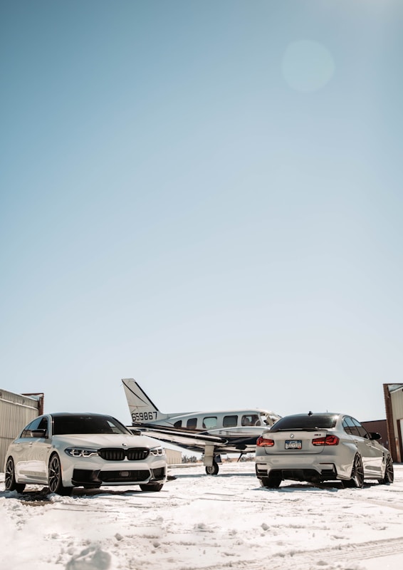 Two luxury cars are parked on a snow-covered surface in front of a small airplane at an airfield. The scene is set under a clear blue sky with hangars visible on either side.